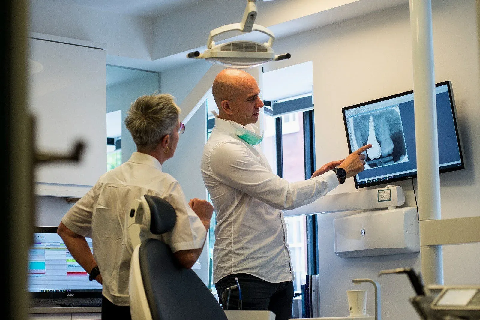 Dentist in a white shirt pointing at a dental X-ray on a wall screen while talking with a woman in a treatment room