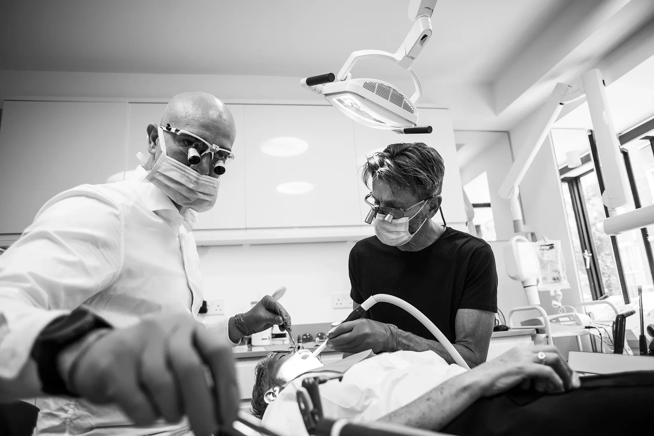 Black and white photo of two masked clinicians wearing loupes treating a patient in a dental chair