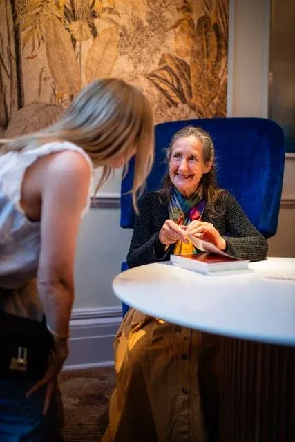 Woman seated at a round table signing a book as another woman leans in to talk to her