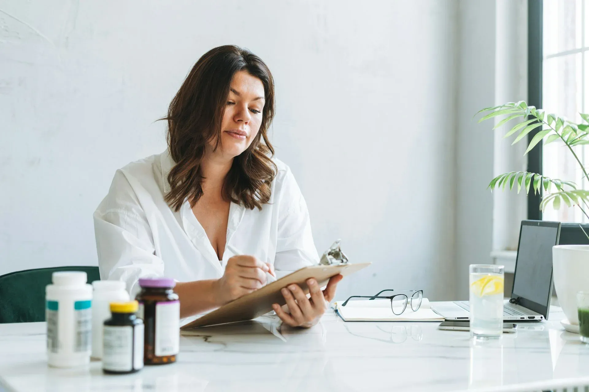 Woman at a white desk writing on a clipboard, with supplement bottles, a laptop and a glass of water