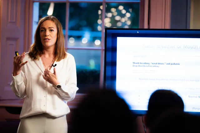Woman speaking to an audience beside a screen showing a slide about nasal breathing and mouth breathing