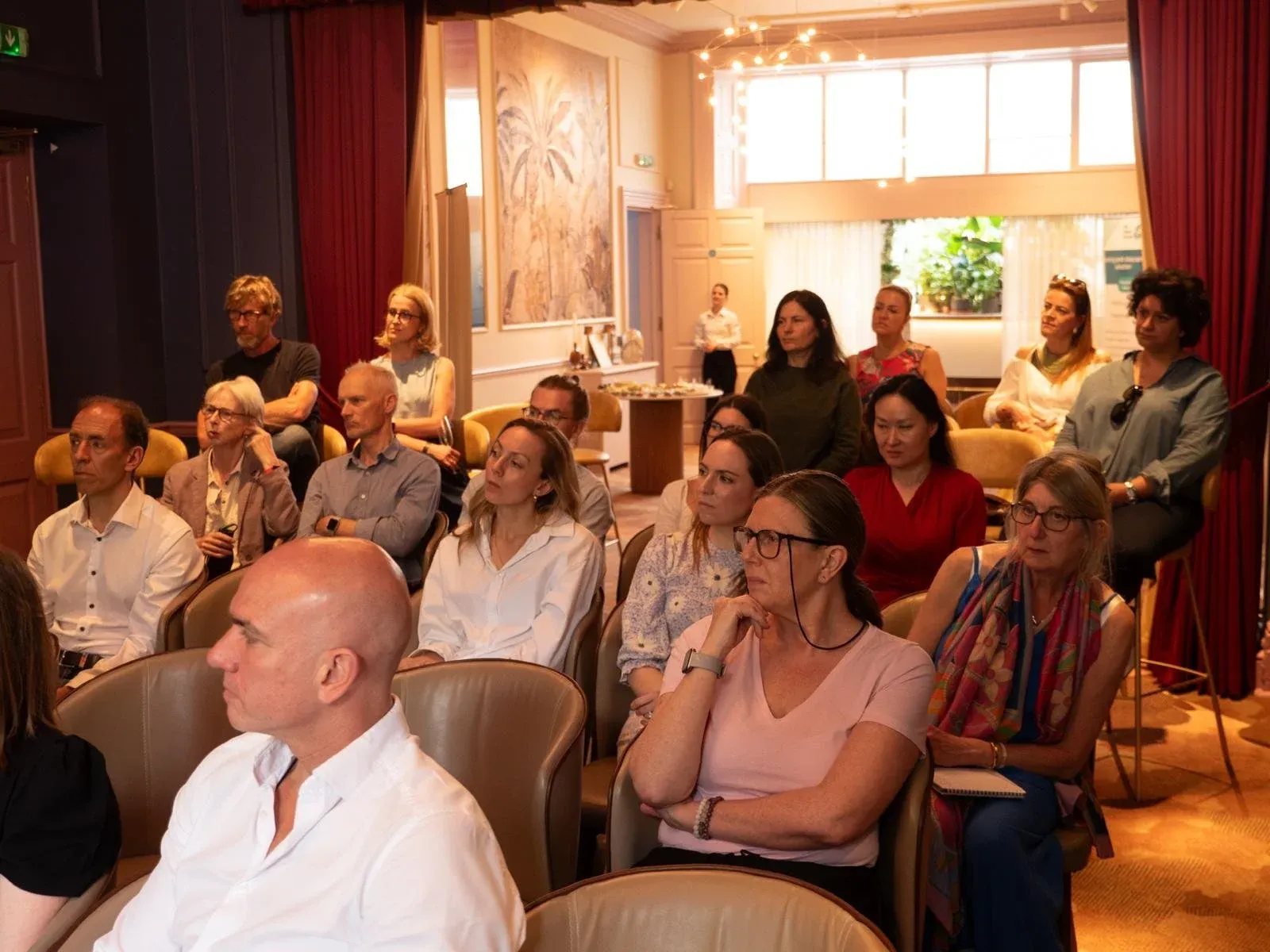 Audience seated in rows in a room with red curtains, listening to a talk
