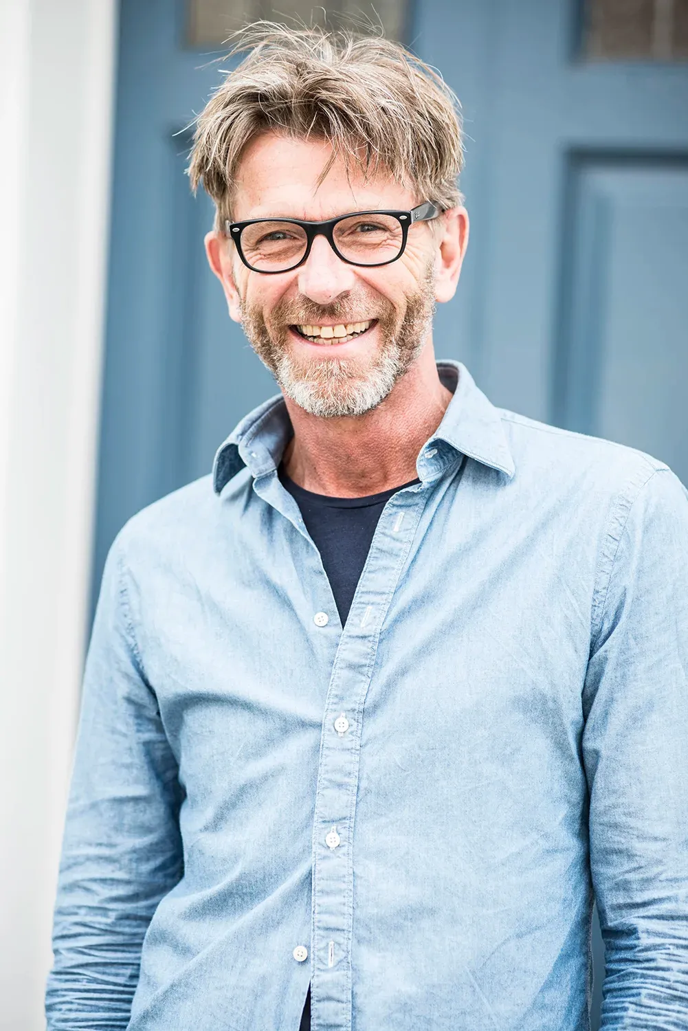 Peter de Jong smiling, in glasses and a light blue shirt, in front of a blue door