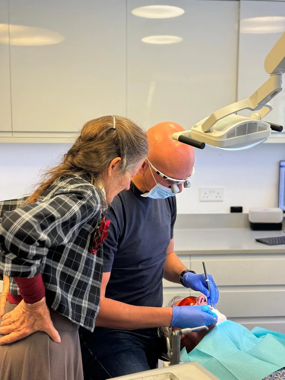 Woman in a checked shirt watching a dentist in a mask treat a patient in a dental chair