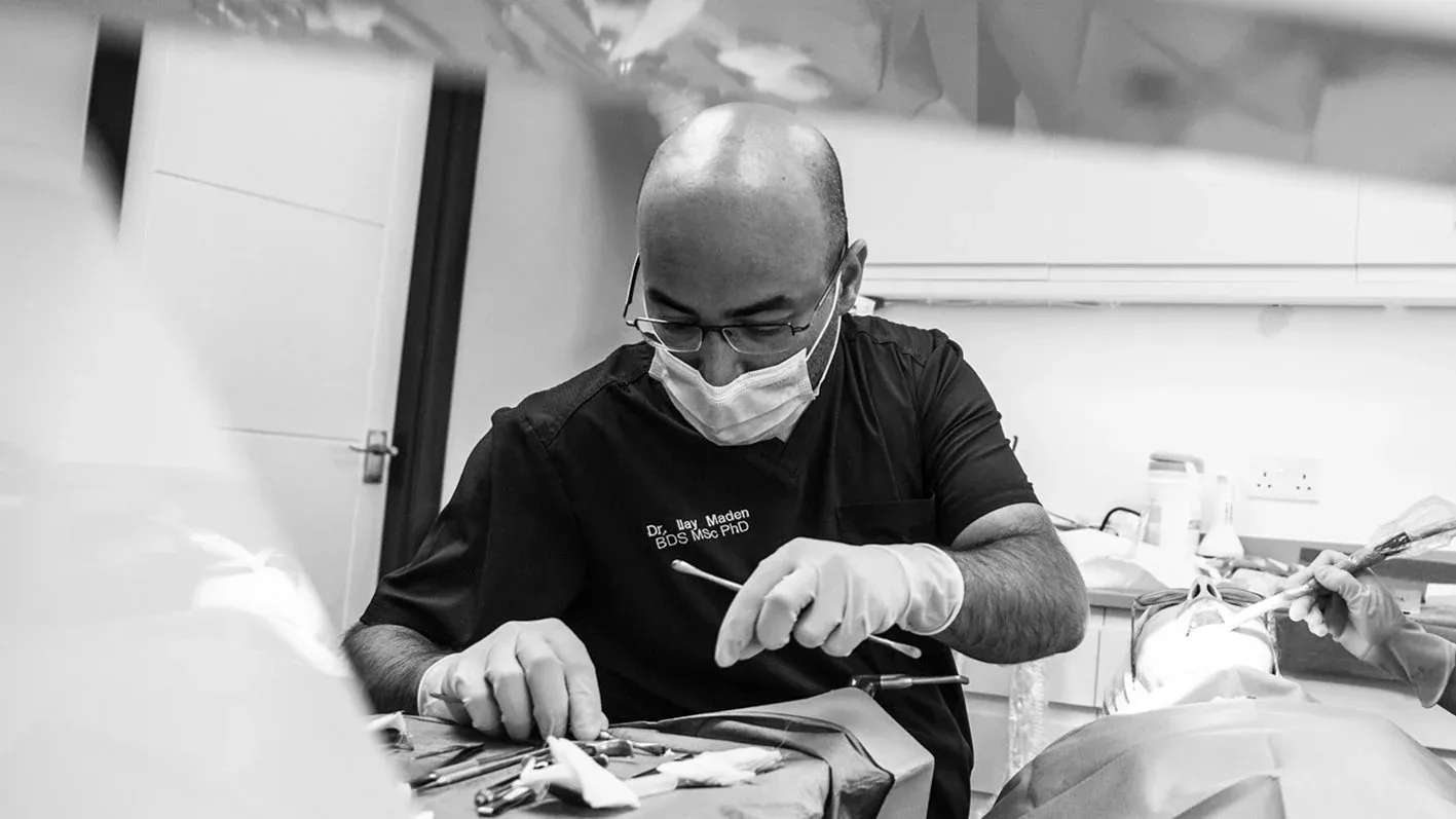 Black and white photo of a masked dentist in dark scrubs holding dental instruments over a treatment tray