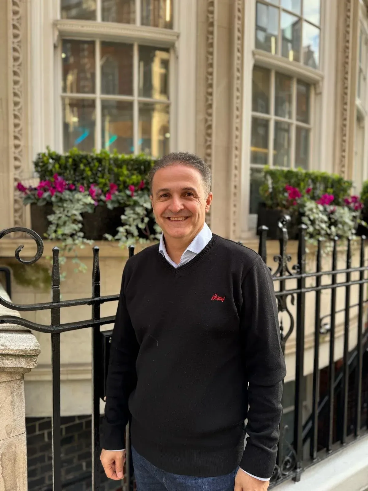 Man in a black jumper smiling beside black railings and window boxes of pink flowers